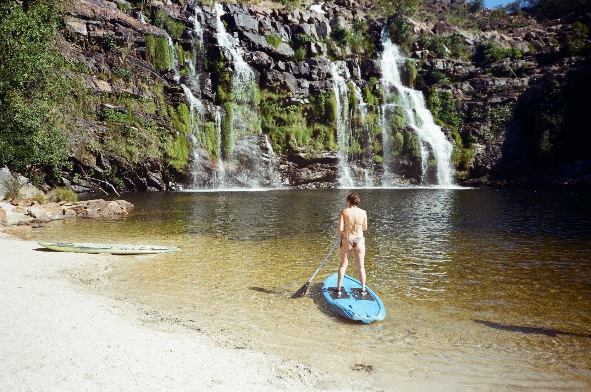 A woman paddleboards on a serene lake near a stunning waterfall, enjoying a sunny outdoor adventure.