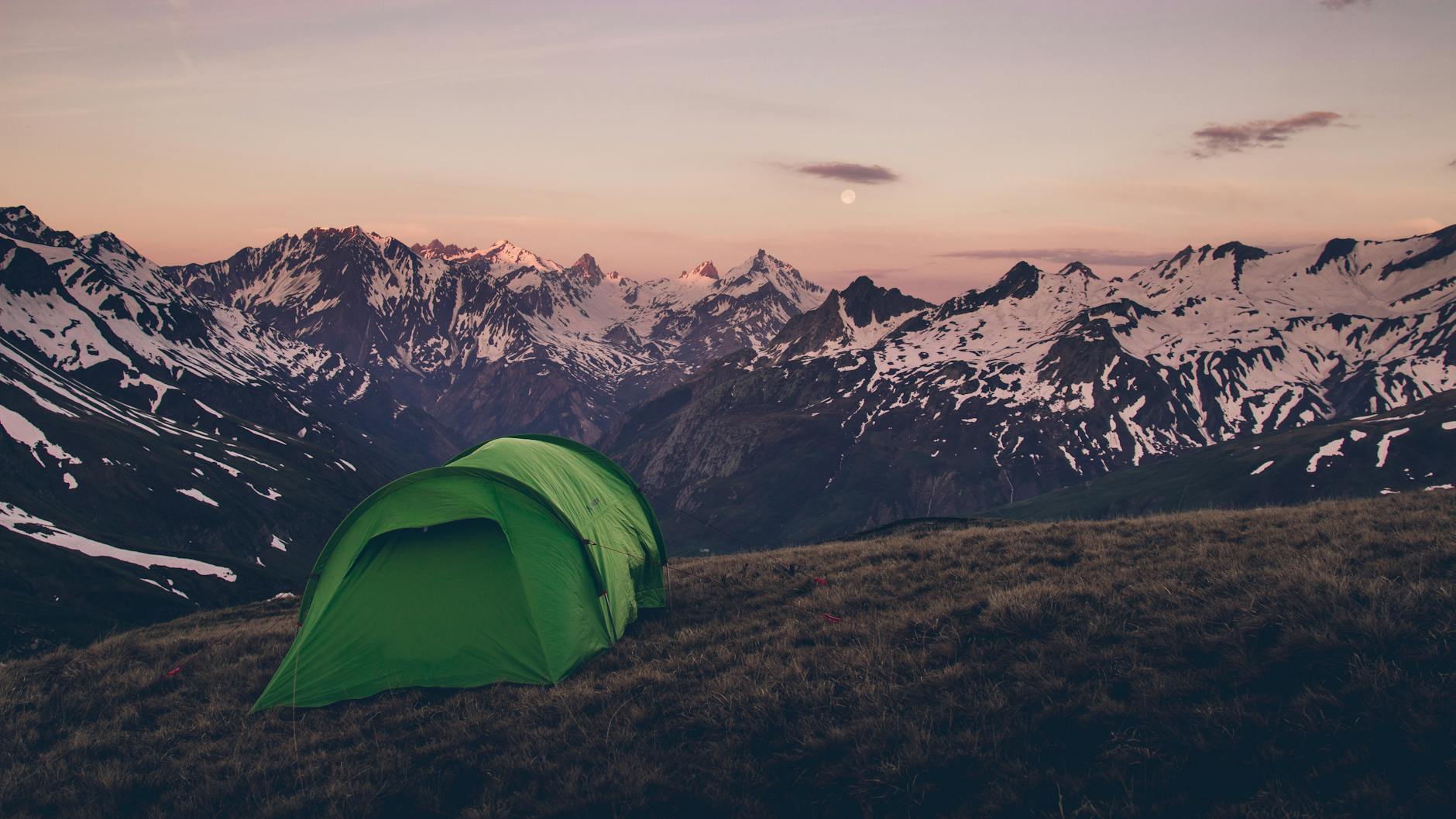 Green tent pitched on a mountain top with stunning snow-capped peaks at dusk.