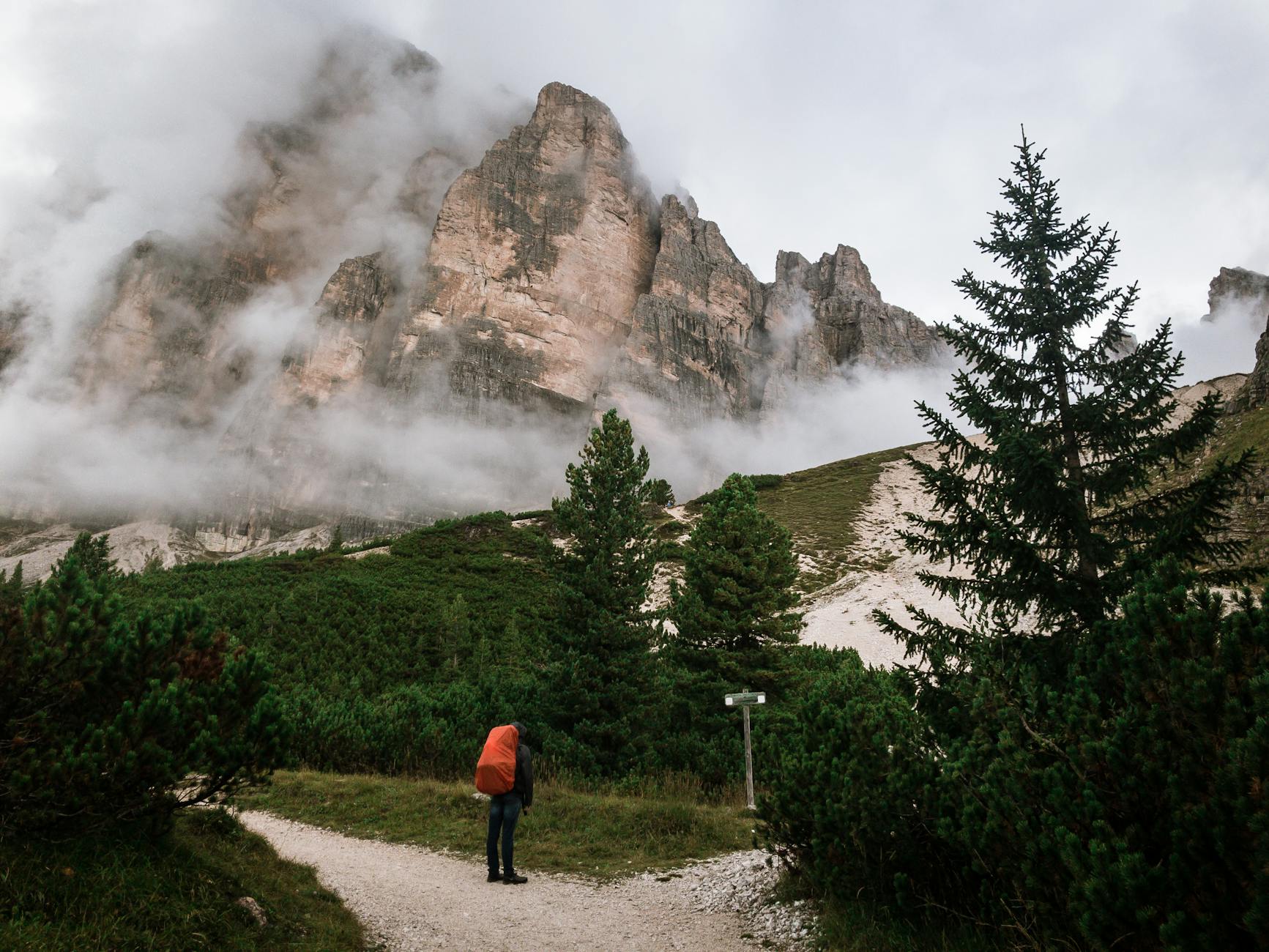 A lone hiker with a red backpack stands on a trail in the misty Dolomites, Cortina d’Ampezzo, Italy.