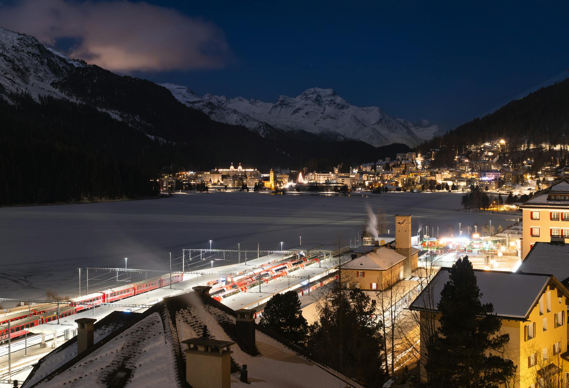 Scenic nighttime view over St. Moritz with illuminated train station and snowy mountains.